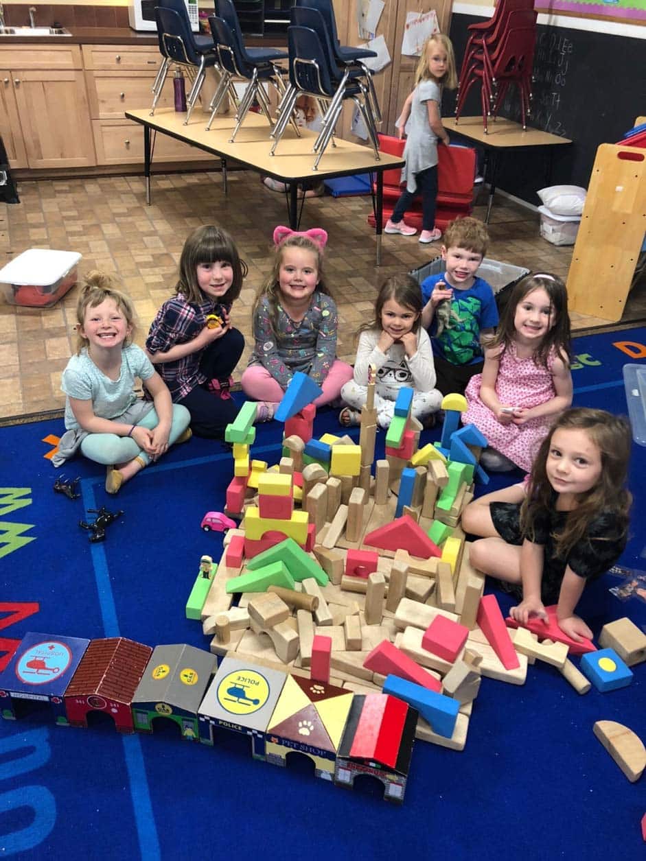 Preschool classmates smiling beside a colorful block structure they built together.
